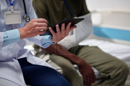 Foto de Primer plano del médico usando tableta digital y hablando con el paciente mientras está sentado en la sala del hospital - Imagen libre de derechos