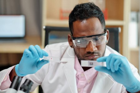 Téléchargez les photos : Jeune homme scientifique sérieux en blouse de laboratoire, gants de protection et lunettes regardant des diapositives lors d'une expérience en laboratoire clinique - en image libre de droit