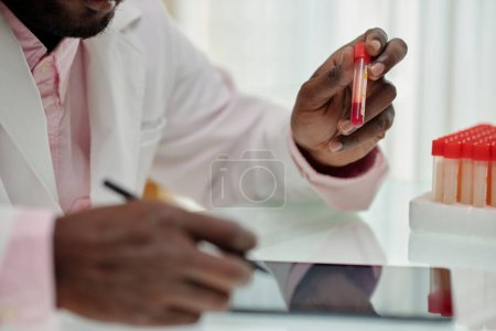 Téléchargez les photos : Main du jeune clinicien afro-américain tenant une fiole avec un échantillon de sang tout en étudiant les caractéristiques du nouveau virus en laboratoire - en image libre de droit