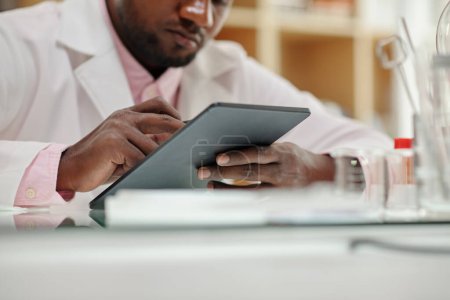 Téléchargez les photos : Gros plan des mains du jeune chercheur afro-américain faisant défiler les informations en ligne à l'écran de la tablette en laboratoire - en image libre de droit