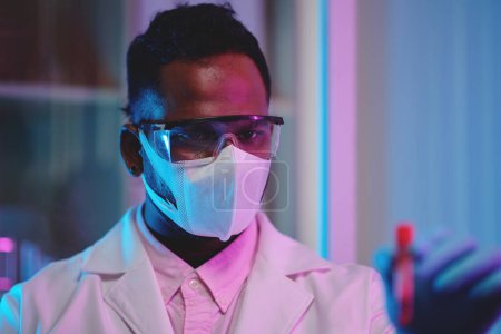Photo for Young serious male chemist in protective mask and eyeglasses looking at sample of new vaccine or medicament during clinical research - Royalty Free Image