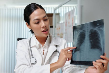 Téléchargez les photos : Jeune radiologiste en blouse de laboratoire commentant la radiographie pulmonaire au patient lors d'une consultation en ligne alors qu'elle était assise dans un cabinet médical - en image libre de droit