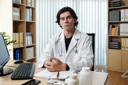 Téléchargez les photos : Jeune télédoc masculin sérieux en blouse de laboratoire assis sur le lieu de travail devant un écran d'ordinateur dans un bureau médical et regardant la caméra - en image libre de droit