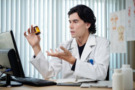 Foto de Clínico varón confiado recomendando nuevas píldoras al paciente en línea en la pantalla del ordenador y explicando por qué es tan eficaz - Imagen libre de derechos