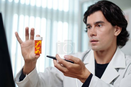Téléchargez les photos : Télédoc confiant avec stylo tenant une petite bouteille avec des pilules tout en les recommandant au patient lors de la consultation en ligne - en image libre de droit