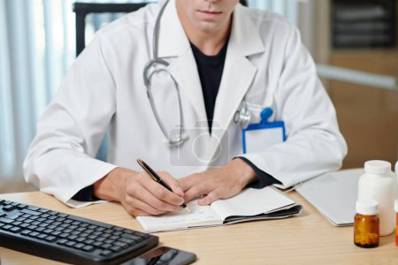 Téléchargez les photos : Gros plan du jeune médecin masculin en blouse de laboratoire prenant des notes sur la carte médicale du patient en ligne ou écrivant des prescriptions pendant la consultation - en image libre de droit