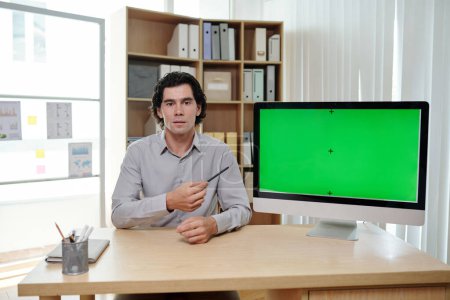 Foto de Joven entrenador de negocios masculino apuntando a la pantalla verde de la computadora mientras está sentado en el lugar de trabajo y haciendo una presentación al público - Imagen libre de derechos