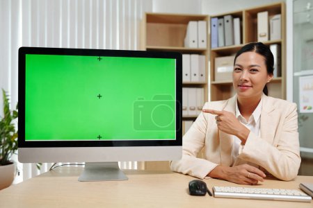 Foto de Joven atractiva morena empresaria apuntando a la pantalla verde de la computadora y mirando a la cámara mientras hace la presentación - Imagen libre de derechos