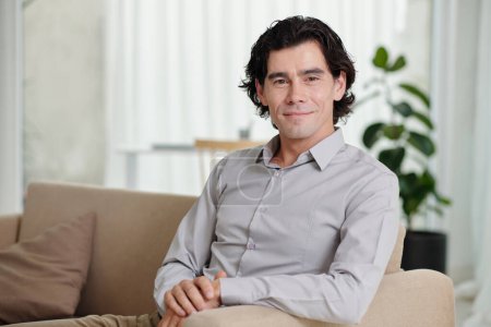 Foto de Joven hombre de negocios sonriente, blogger o psicólogo con camisa azul claro sentado en el sofá en el entorno doméstico y mirando a la cámara - Imagen libre de derechos