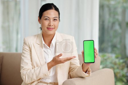 Photo for Happy young brunette businesswoman presenting new mobile application while holding smartphone with green screen and looking at camera - Royalty Free Image