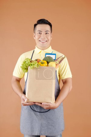 Foto de Trabajador de supermercado asiático sonriente sosteniendo bolsa de papel llena de comestibles frescos, frutas y verduras - Imagen libre de derechos