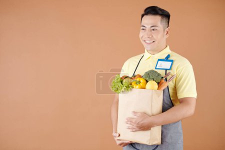 Foto de Trabajador de tienda oscura asiático feliz que lleva la bolsa de productos frescos recogidos para la entrega al cliente - Imagen libre de derechos