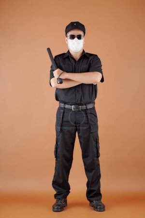 Foto de Retrato del guardia de seguridad confiado en máscara médica sosteniendo bastón, cruzando brazos y mirando a la cámara - Imagen libre de derechos