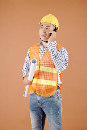 Photo for Portrait of serious construction engineer with blueprint in hand talking on phone with contractor - Royalty Free Image