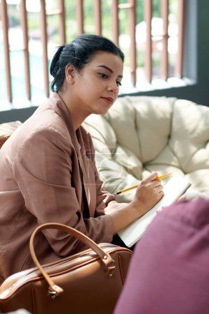 Téléchargez les photos : Jeune femme d'affaires confiante prenant des notes dans le planificateur lorsqu'elle parle à un collègue à la réunion - en image libre de droit