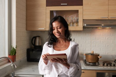 Téléchargez les photos : Positif jeune femme debout dans la cuisine et la lecture des nouvelles sur les médias soacial via l'application sur tablette - en image libre de droit