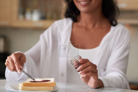 Photo for Woman speading homemade jelly from small jar on toast when making breakfast in the morning - Royalty Free Image