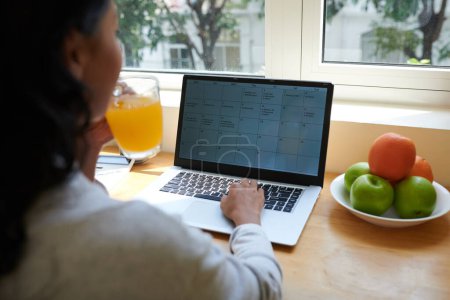 Téléchargez les photos : Jeune femme buvant une tasse de jus et vérifiant le calendrier et le calendrier sur l'écran d'ordinateur portable - en image libre de droit