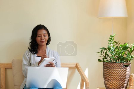Téléchargez les photos : Jeune femme créative assise dans un fauteuil à la maison, travaillant sur un ordinateur portable et écrivant des idées dans un planificateur - en image libre de droit