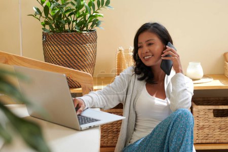 Téléchargez les photos : Joyeux jeune femme travaillant sur ordinateur portable et parlant au téléphone avec un collègue - en image libre de droit