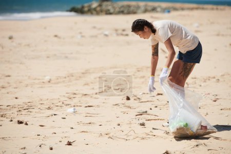 Photo for Young woman cleaning up sea beach, collecting trash into plastic bag - Royalty Free Image