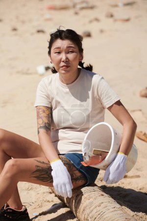 Téléchargez les photos : Portrait d'une bénévole se reposant sur un tronc d'arbre sur la plage après avoir ramassé des ordures dans un seau en plastique - en image libre de droit