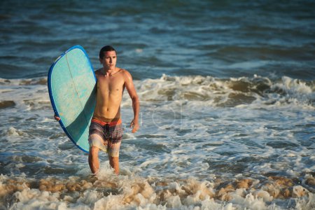 Téléchargez les photos : Jeune homme sortant de l'océan après avoir surfé dans un rayon de soleil couchant - en image libre de droit