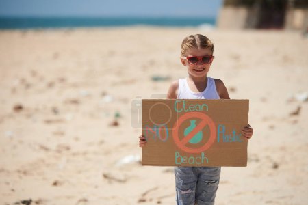 Téléchargez les photos : Joyeux petite fille en lunettes de soleil tenant une plaque en carton essayant d'organiser le nettoyage de la plage - en image libre de droit