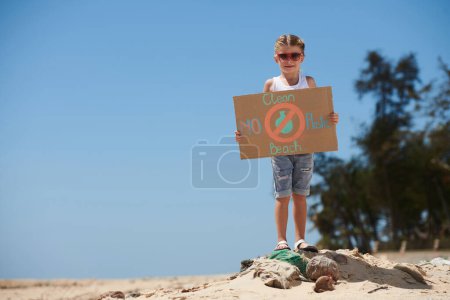 Téléchargez les photos : Petite fille sans plastique et nettoyer la plaque de la plage debout sur un tas de déchets sur la plage - en image libre de droit