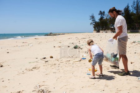 Foto de Padre enseñando a su hijita a recoger desechos plásticos en la costa del océano arenoso para salvar el futuro - Imagen libre de derechos