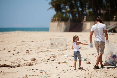 Téléchargez les photos : Père et fille marchant sur une plage sale et ramassant des déchets plastiques dans un gros sac le jour de la Terre - en image libre de droit