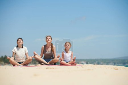 Photo for Little girl and two women meditating in lotus position on sandy beach and enjoying sea breeze - Royalty Free Image