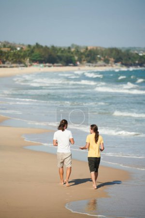 Téléchargez les photos : Jeunes hommes aux cheveux longs parlant et marchant sur la plage par une journée ensoleillée - en image libre de droit