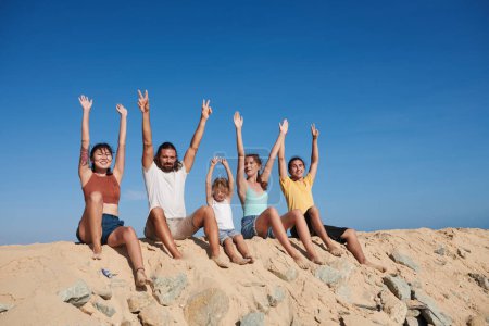Photo for Group of joyful people sitting on beach, raising arms, enjoying sunrays and sea breeze - Royalty Free Image