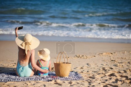 Téléchargez les photos : Jeune femme prenant selfie avec petite fille quand ils sont assis sur la plage dans des chapeaux de paille - en image libre de droit