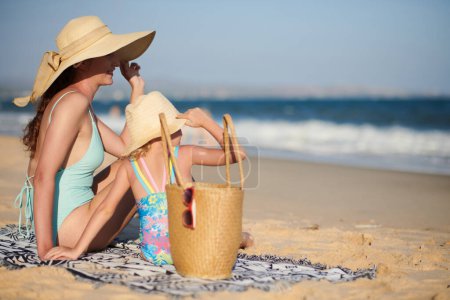 Téléchargez les photos : Mère et fille heureuses assises sur une plage de sable et regardant les vagues de mer - en image libre de droit
