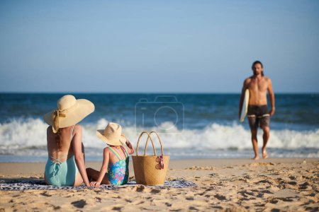 Foto de Mujer con sombrero de paja grande y su hija mirando al marido regresar después de surfear en el mar - Imagen libre de derechos