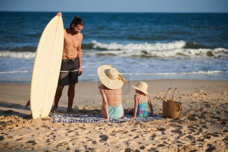 Téléchargez les photos : Souriant jeune homme parlant à sa femme et sa petite fille après avoir surfé en mer - en image libre de droit