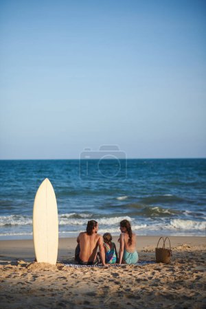 Téléchargez les photos : Famille de trois personnes assise sur une couverture sur la plage et regardant les vagues de la mer - en image libre de droit