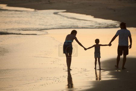 Foto de Familia feliz de tres personas cogidas de la mano al caminar en la playa de arena al atardecer - Imagen libre de derechos