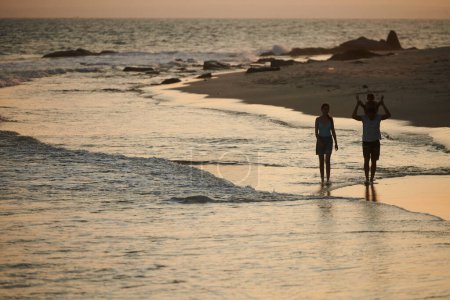 Téléchargez les photos : Homme donnant tour de dos à sa fille lors de la promenade sur la plage avec la famille au coucher du soleil - en image libre de droit