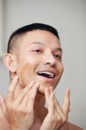 Téléchargez les photos : Appliquer jeune homme appliquant un masque d'argile rouge sur le menton pour nettoyer en profondeur les pores - en image libre de droit