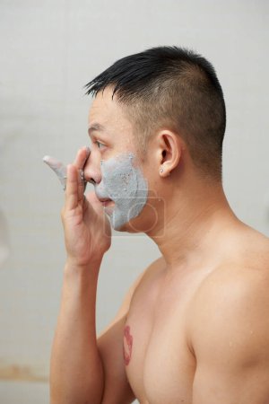 Téléchargez les photos : Jeune homme torse nu regardant miroir de salle de bains lors de l'application profonde pore nettoyage masque d'argile - en image libre de droit