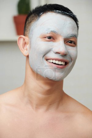 Téléchargez les photos : Portrait de jeune homme happly avec masque en argile kaolin sur le visage debout dans la salle de bain - en image libre de droit
