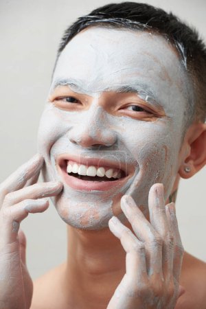 Téléchargez les photos : Portrait d'un jeune homme joyeux appliquant un masque d'argile à resserrer les pores sur le visage - en image libre de droit