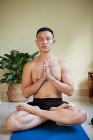 Téléchargez les photos : Portrait de jeune homme sérieux assis en position lotus dans un tapis de yoga et méditant les yeux fermés - en image libre de droit