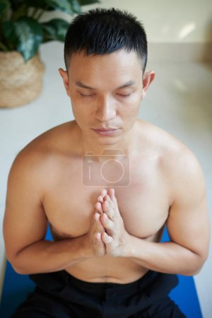 Foto de Tranquilo joven sin camisa serio manteniendo las manos en gesto mudra al meditar en casa por la mañana - Imagen libre de derechos