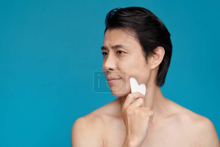 Photo for Man massaging his face with gua sha tool, isolated on blue - Royalty Free Image