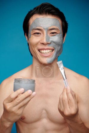 Téléchargez les photos : Portrait de l'homme en forme excité appliquant masque de charbon sur son visage - en image libre de droit