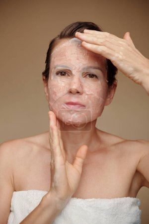 Foto de Mujer madura aplicando máscara de sábana de su cara después de la ducha de la mañana - Imagen libre de derechos